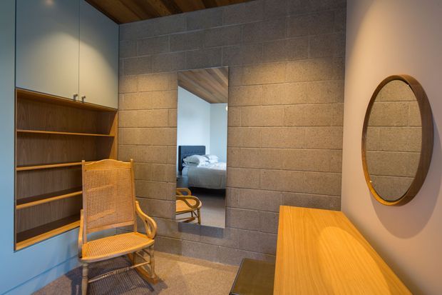 Warm, compact bedroom dressing or wardrobe area with exposed grey masonry block walls, built-in timber shelving and cabinetry, a cane chair, timber ceiling, and a narrow full-height mirror reflecting a bed beyond. A curved timber console and round framed wall mirror add to the natural, mid-century-inspired interior.