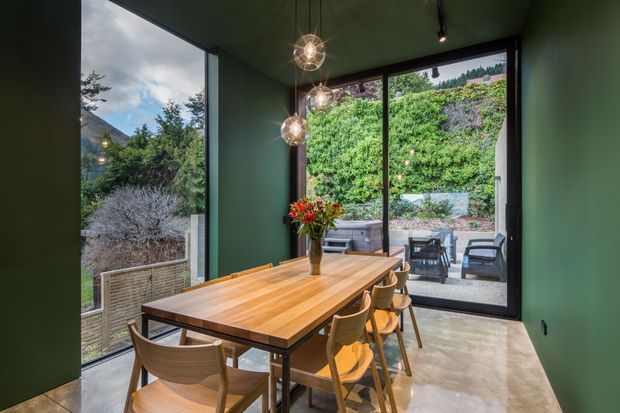 A compact, contemporary dining room with a long timber dining table and matching wooden chairs, enclosed by expansive floor-to-ceiling glazing. Dark green walls and ceiling create a rich, moody backdrop, while spherical pendant lights hang above the table. The glazing opens views to a landscaped outdoor seating area and surrounding vegetation.