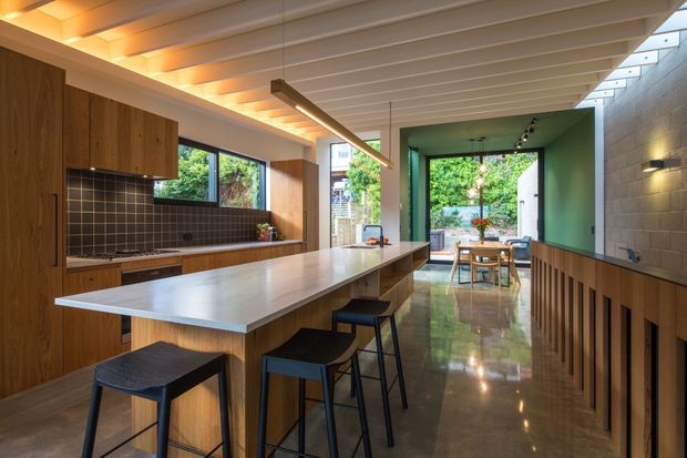 A contemporary open-plan kitchen and dining space with a long central island, timber cabinetry, dark square-tile splashback, and integrated bar seating. The room features polished concrete flooring, a ribbed timber ceiling with warm recessed lighting, a linear pendant, and a green-painted dining alcove opening to a leafy outdoor area.
