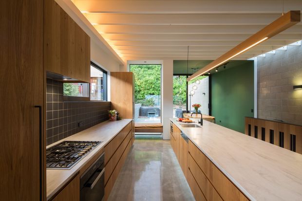 A contemporary galley-style kitchen with a large central island, warm timber cabinetry, pale stone benchtops, dark tiled splashback, polished concrete flooring, and a green feature wall. The space has a white exposed-beam ceiling, integrated linear pendant lighting, and glazing opening toward a leafy outdoor area.