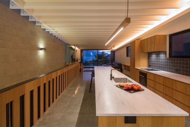 Long, narrow contemporary kitchen with a large pale stone island, integrated sink and seating, warm timber cabinetry, dark tiled splashback, polished concrete floor, and a linear pendant light. A timber-slatted balustrade borders the walkway beside a textured masonry wall, with glazing at the far end overlooking the landscape.