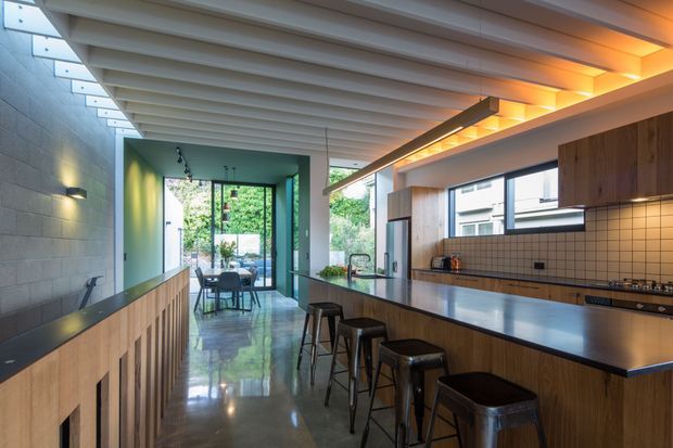 A spacious contemporary kitchen with a long island and bar seating, warm timber cabinetry, dark benchtops, and a tiled splashback. The room features an exposed ribbed ceiling with integrated linear lighting, polished concrete flooring, clerestory-style windows, and a glazed opening leading to a dining area and greenery beyond.