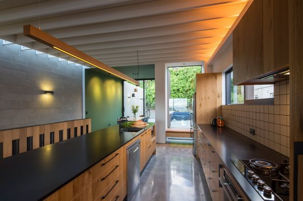 A narrow, contemporary galley kitchen with a central timber island and black benchtop, matching timber cabinetry along the right wall, and a tiled splashback. The kitchen features a polished concrete floor, exposed pale ceiling battens, integrated warm strip lighting, and a glazed door at the rear opening to greenery outside.
