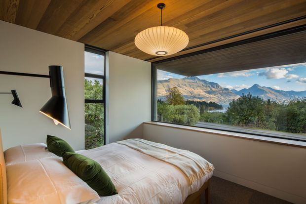 A warm, contemporary bedroom with a low platform bed dressed in white bedding and green cushions. The room features a wood-lined ceiling, large black-framed picture windows, a sculptural wall-mounted bedside light, and expansive views of a lake and mountains beyond.