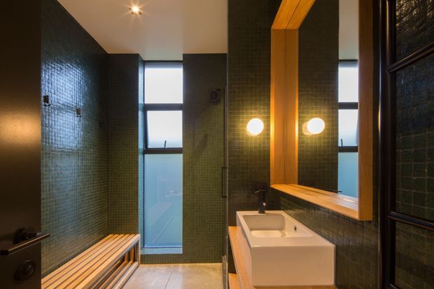 A compact contemporary bathroom with dark green small-format tiled walls, a white vessel basin on timber cabinetry, and a large timber-framed mirror with round wall lights. A glazed shower enclosure and timber slatted bench are visible, with warm lighting creating a refined, moody atmosphere.