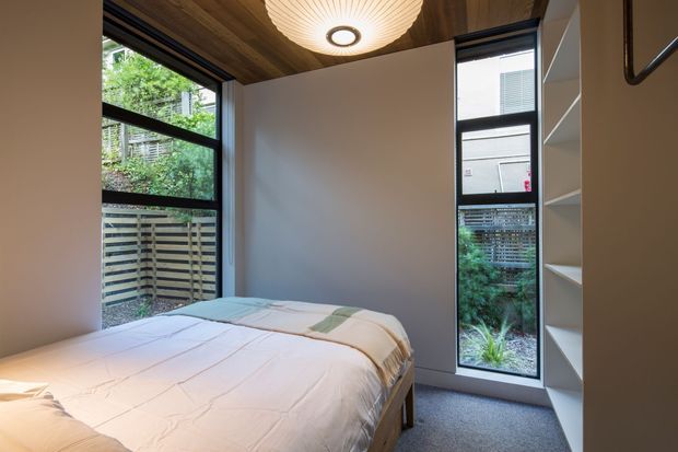 Compact contemporary bedroom with a double bed, neutral bedding, built-in open shelving, tall black-framed windows, and a warm timber ceiling. A sculptural circular ceiling light adds a soft focal point, while greenery and a slatted fence are visible outside.