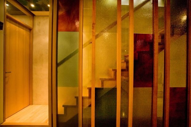 Warmly lit interior view of a compact stairway enclosed by translucent textured glass panels set within timber framing. Timber doors, light timber flooring, exposed timber ceiling beams, and bold translucent coloured glass create a dramatic layered partition. The staircase is visible behind the glazing, with the camera positioned in an adjoining hallway or entry area.