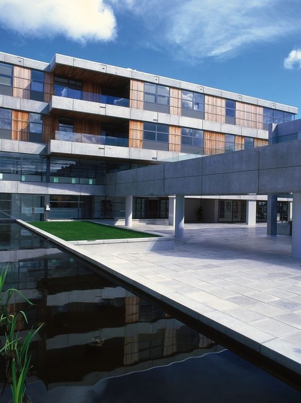 Exterior architectural view of a contemporary multi-storey building arranged around a landscaped courtyard and reflecting water feature. The facade combines exposed concrete bands and columns, extensive dark-framed glazing, and vertical timber cladding panels. Deep balconies, projecting volumes, and a covered open-air ground level create a strong geometric composition. Bright blue sky and sunlight emphasize the crisp modern materials and reflections in the pool.