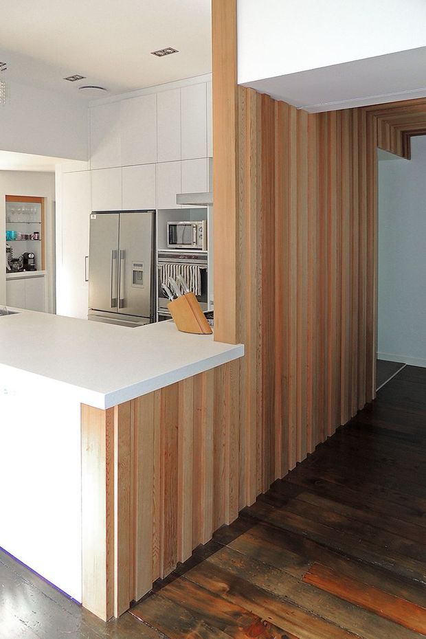 Interior view of a contemporary kitchen opening onto a dark timber-floored hallway. The kitchen features extensive white built-in cabinetry, a large white island benchtop, stainless-steel appliances, and a timber-clad island base. Vertical natural timber battens continue along the adjacent wall, creating a strong feature surface and warm contrast with the white joinery and dark flooring.