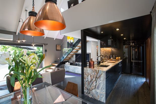 Open-plan contemporary kitchen and dining area with a dramatic black kitchen fit-out, veined stone island, dark timber flooring, and copper pendant lights. A living area and staircase are visible beyond the kitchen, with abundant daylight entering through large windows.