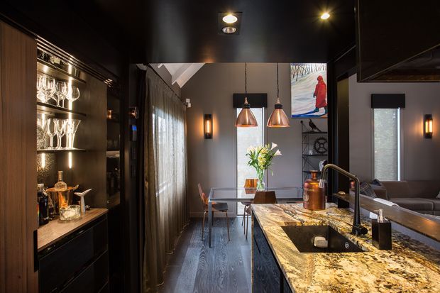 A dramatic, dark-toned contemporary kitchen and dining area. The kitchen features a large marble-patterned island with an undermount sink, tall black tap, dark cabinetry, integrated appliances, and a backlit display cabinet for glassware and spirits. Copper pendant lights hang above the dining table, while a grey sofa, artwork, shelving, and warm wall sconces extend into the adjoining living area.