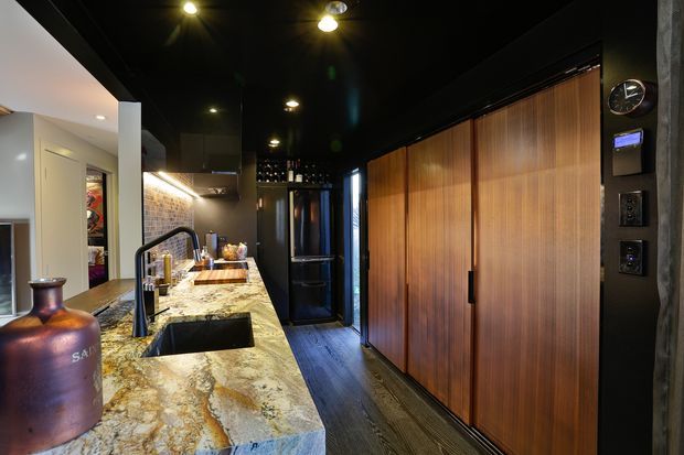 A dramatic, narrow contemporary kitchen with a long natural-stone island and integrated sink, dark cabinetry and ceiling, warm timber veneer storage doors, and a dark wood-look floor. Recessed lighting and under-cabinet illumination create a moody, high-contrast atmosphere, while a large decorative vessel sits on the island.