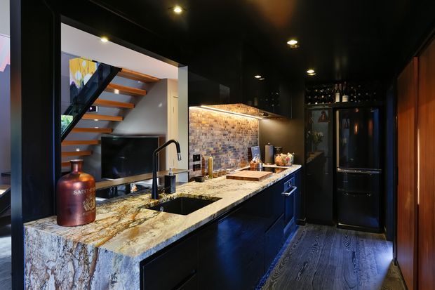 A compact, dramatic kitchen with a black ceiling, dark cabinetry and appliances, a heavily patterned stone waterfall island and matching benchtop, and a textured exposed-brick splashback. A black undermount sink and tap sit in the island, while a staircase and living area are visible beyond the opening. Integrated display shelving and a timber-toned vertical wall panel add warmth.