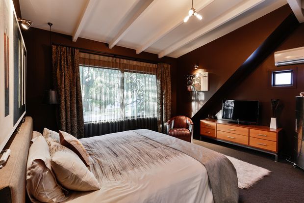 A warm, moody bedroom with a large upholstered bed, layered neutral bedding, dark brown walls, and heavy patterned curtains. White exposed ceiling beams contrast with the dark interior, while a timber mid-century-style dresser supports a television. A leather armchair, framed artwork, small high-set window, and diagonal black structural beam complete the room.