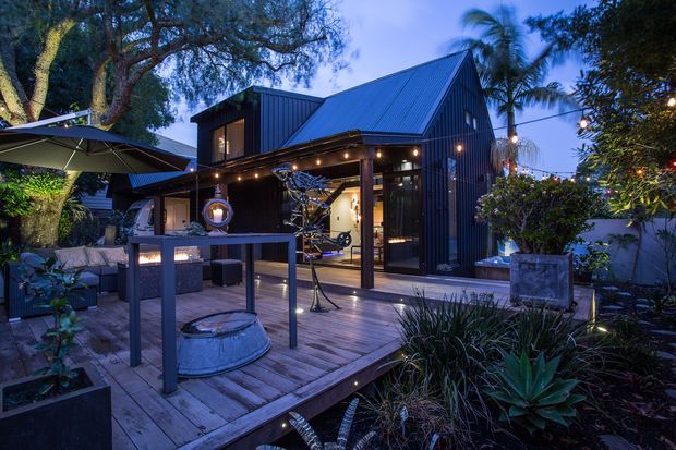 Twilight exterior view of a compact dark-clad gabled pavilion or studio opening onto a timber deck. The building features a steep pitched metal roof, large glazed doors, covered outdoor entertaining space with festoon lighting, a bar-height table, outdoor furniture, and lush subtropical planting. A glimpse inside shows exercise equipment, suggesting a home gym or studio.