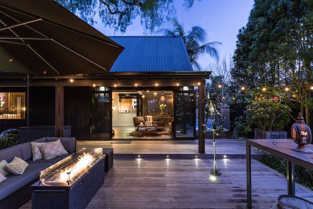 Twilight view of a refined outdoor deck and entertaining area adjoining a dark-clad pavilion or house. The scene features timber decking, a large outdoor sectional sofa, a linear fire table, an umbrella, a dining table, festoon lighting, planting, and wide glazed doors opening to a warmly lit interior living space. A pitched corrugated metal roof and dark timber-style exterior cladding are visible.