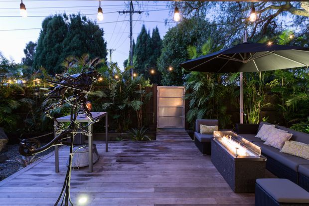 A warmly lit outdoor entertaining area on a timber deck, surrounded by dense tropical and mature garden planting. The space includes dark outdoor lounge seating, a large cantilevered umbrella, a rectangular fire feature or table, festoon lighting, and a bicycle displayed near the deck entrance.