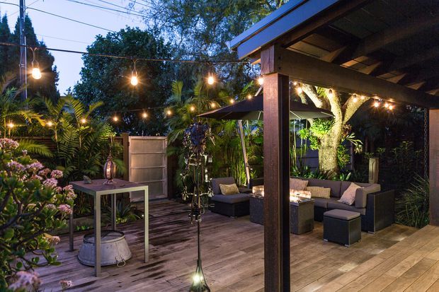 A warmly lit covered outdoor entertaining area with timber decking, a timber-framed roof structure, built-in or freestanding lounge seating, and abundant tropical garden planting. String lights create an inviting evening atmosphere, while a small outdoor table and decorative lighting add to the relaxed courtyard setting.