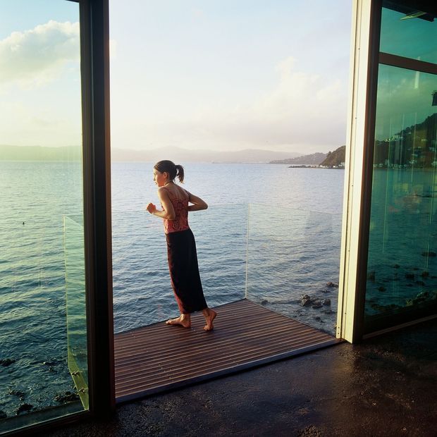 A modern interior opens directly onto a narrow timber deck over a calm body of water. A full-height glazed opening with dark frames and a glass balustrade creates a seamless indoor–outdoor connection, with a person standing on the deck and a distant wooded shoreline beyond. The space has a dark, polished-looking floor and strong coastal outlook.