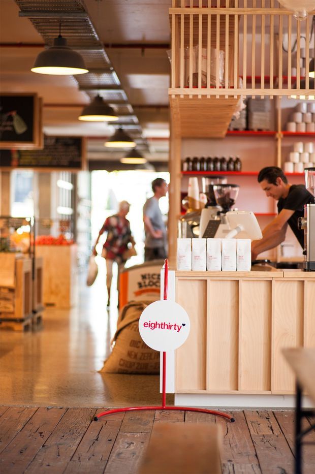 A warmly lit specialty coffee shop interior with a timber service counter, espresso equipment, shelves of coffee products, hanging industrial-style pendant lights, timber flooring, and a partially screened overhead mezzanine or storage structure. Staff and customers are visible in the background, with coffee sacks near the counter and a branded circular sign in the foreground.