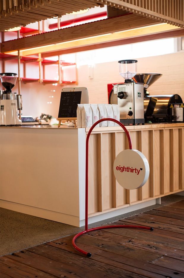 A warmly lit specialty coffee counter with commercial espresso and coffee-grinding equipment, coffee bags, and a small menu board. The counter combines pale smooth cabinetry with vertical light timber battens, while exposed timber slats form a layered ceiling feature. A red circular branded sign and red shelving add strong accents. The space has a contemporary hospitality fit-out with an industrial coffee-shop character.