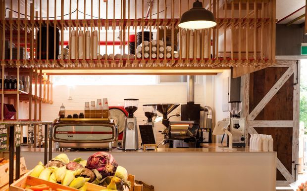 A warmly lit commercial café or coffee shop interior, viewed toward a service counter with multiple espresso grinders and coffee equipment. A large refrigerated display case of fresh produce occupies the foreground, while shelves and a suspended timber slat storage/display structure hold bottles, jars, and cylindrical containers overhead. The space combines white cabinetry and counters with exposed timber framing, a dark pendant light, and a large rustic barn-style timber door at right.
