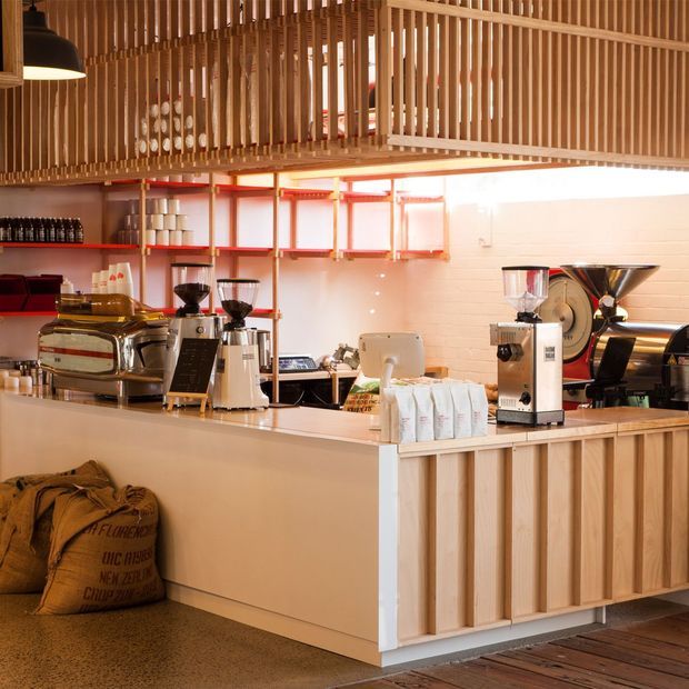 Warmly lit coffee shop interior viewed from the customer side of a timber-clad service counter. The space features espresso machines and coffee grinders, open red shelving with cups and bottles, a white tiled or brick-patterned rear wall, and a prominent suspended timber slat ceiling and upper screen. Burlap coffee sacks sit beneath the counter, with polished concrete and timber flooring visible.