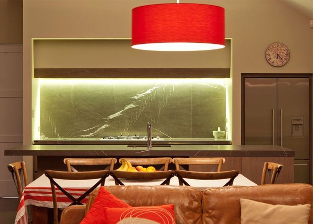 Warm, stylish open-plan kitchen and dining area viewed from a living space. The kitchen features a long dark island with sink, a dramatic illuminated green-toned stone backsplash, dark wood cabinetry, and integrated appliances. A large red pendant light hangs over the island, while a timber dining table with leather and timber chairs sits in the foreground, partially behind a brown leather sofa. The room has layered warm lighting and a rich, earthy palette.