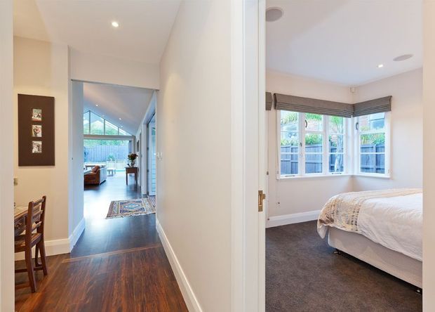 Interior view looking along a hallway with dark timber flooring, opening to a bright living area with angled glazing at the far end. A bedroom is visible to the right through an open doorway, featuring a white upholstered bed, grey carpet, white walls, and a bay-style corner window with woven Roman blinds. A small dining area and wall-mounted artwork are visible along the hallway.