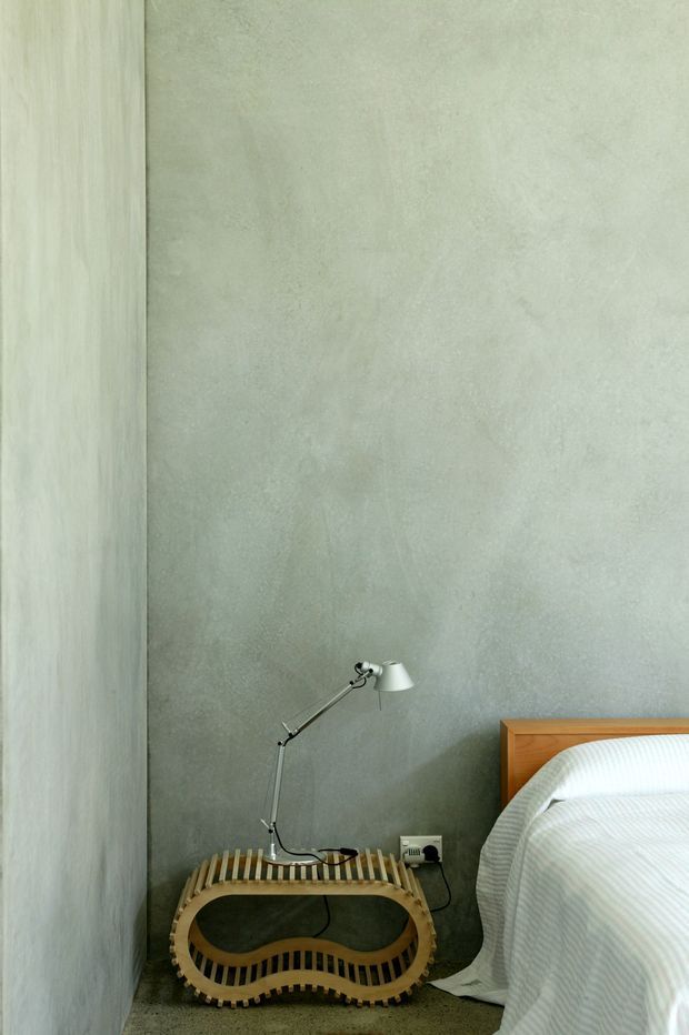 A close-up view of a compact bedroom corner, featuring a bed with a warm timber headboard and white striped bedding. Beside it is a sculptural curved timber side table with a modern adjustable metal desk lamp. The walls have a mottled pale grey-green plaster or concrete-like finish, creating a quiet, minimalist atmosphere.