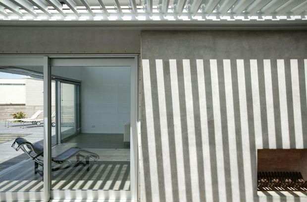 Modern architectural exterior or semi-outdoor terrace showing a raw grey concrete wall dramatically patterned with vertical shadows from a slatted overhead canopy. Large sliding glass doors reveal a minimalist interior with timber flooring and lounge chairs, while a dark timber bench sits against the wall. The composition emphasizes strong geometric lines, daylight, and shadow play.