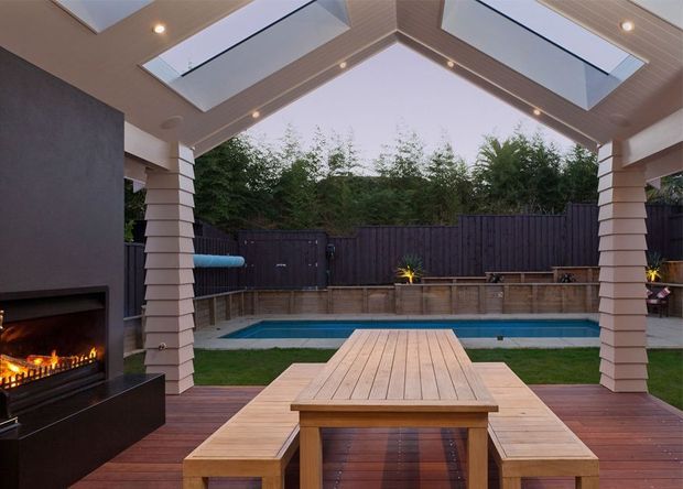 Covered outdoor entertaining area with a timber dining table and built-in bench seating on a timber deck, opening onto a rectangular swimming pool and landscaped courtyard. The space features a large outdoor fireplace, timber-clad pitched roof with multiple skylights, recessed lighting, and pale horizontal cladding around the supporting columns.