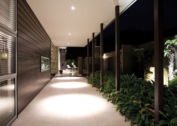 A covered, softly lit architectural walkway beside a contemporary building. The corridor features large pale tiled flooring, dark horizontal wall cladding on the left, full-height dark-framed glazing and screening on the right, recessed ceiling lights, and lush planted beds along the edge. A seating area is visible at the far end.