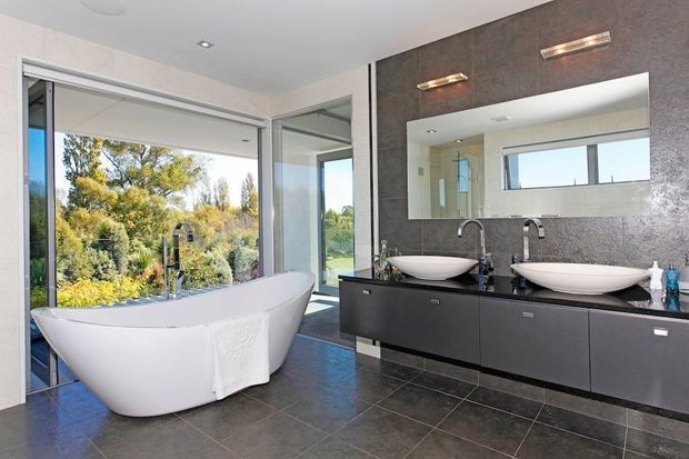 A spacious contemporary bathroom with a freestanding white soaking bathtub beside full-height glazing, overlooking landscaped greenery. A long dark floating vanity has two white vessel basins, wall-mounted chrome taps, a large rectangular mirror, and slim horizontal wall lights. Dark tiled flooring and a dark textured feature wall create a strong modern contrast with the pale walls and white fixtures.