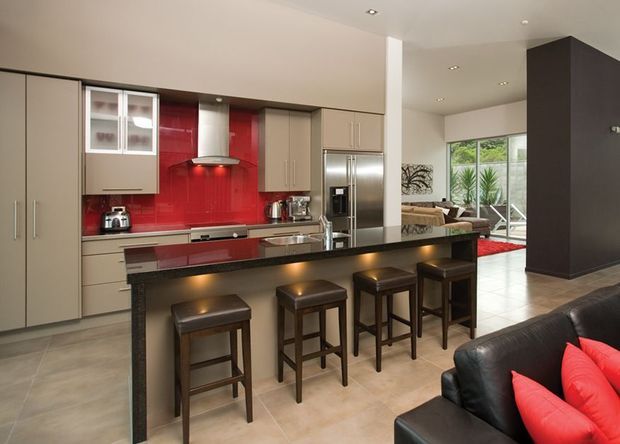Open-plan contemporary kitchen and living area with extensive taupe cabinetry, a glossy red splashback, stainless-steel appliances, a dark stone-look island with four bar stools, and pale tiled flooring. A black leather sofa with red cushions is visible in the adjoining living space, which opens toward a bright glazed outdoor area.