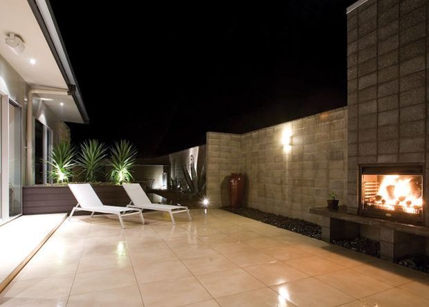 A spacious outdoor courtyard or terrace at night, paved with large light-coloured tiles and enclosed by rendered concrete block walls. Two white lounge chairs sit beside a raised planter with tropical plants, while a built-in fireplace creates a warm focal point on the right. Glazed doors and a covered overhang connect the terrace to the house.