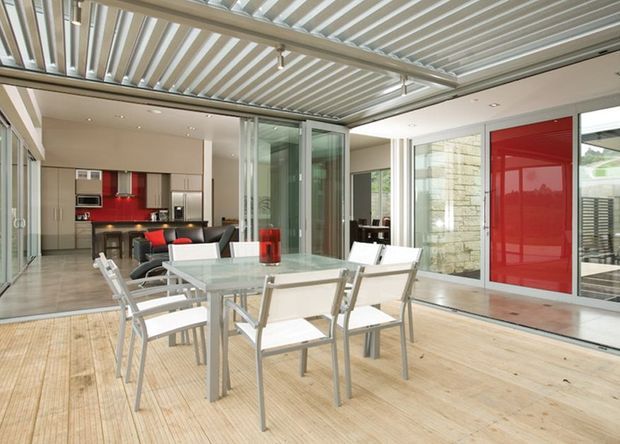 Contemporary indoor-outdoor entertaining area with a timber deck, glass dining table and white outdoor dining chairs beneath a louvred roof. The space opens into a modern kitchen and living area through extensive sliding glazing, with a prominent red feature wall and door adding contrast.