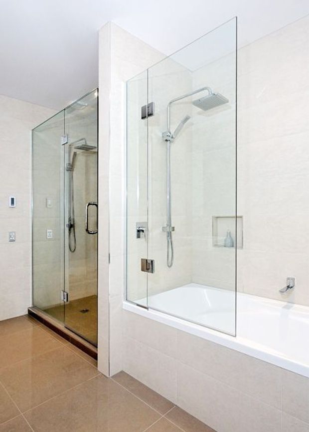 Bright contemporary bathroom featuring a glass-enclosed walk-in shower beside a white bathtub with integrated shower fittings. The space has large light wall tiles, warm taupe floor tiles, chrome fixtures, recessed shelving, and frameless glass panels.