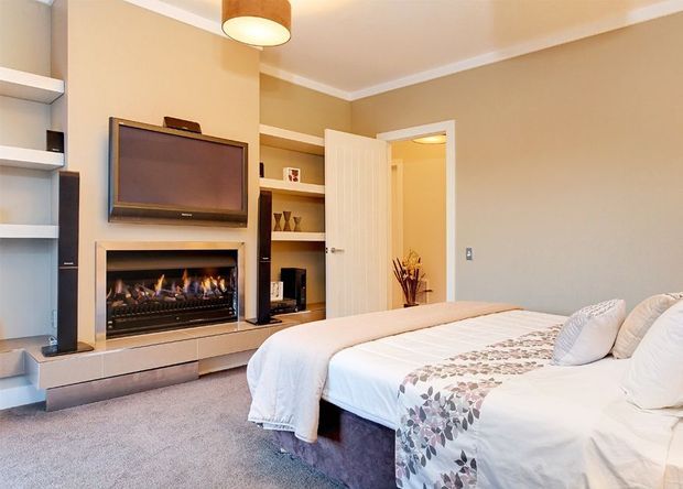 A bright, warmly styled bedroom with a large bed dressed in white bedding and a patterned runner, plush carpet, neutral walls, and built-in shelving. A contemporary built-in entertainment and feature wall includes a wall-mounted television above a linear gas fireplace, with an open door leading to an adjoining space.