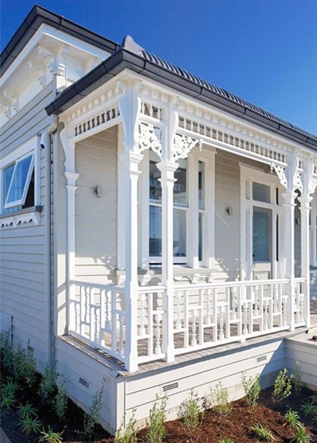 A bright exterior view of a restored or newly built ornate white timber house, featuring a covered front veranda with decorative fretwork, turned posts, balustrades, brackets, and horizontal weatherboard cladding. The roof has dark metal roofing with a prominent gable-like edge, while white-framed windows and a glazed entry door open onto the veranda. Low landscaping borders the base.
