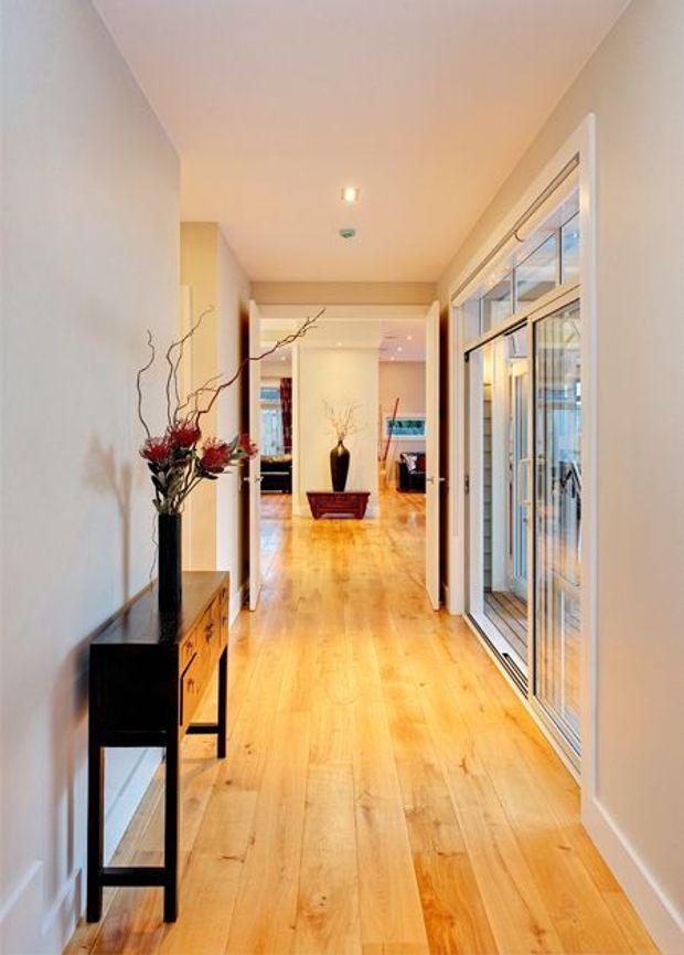 A bright residential hallway with warm timber flooring, pale neutral walls, recessed ceiling lighting, and large glazed sliding doors or windows along the right side. A narrow dark console with drawers and decorative vases occupies the left wall, while the corridor opens toward a furnished living area in the distance.