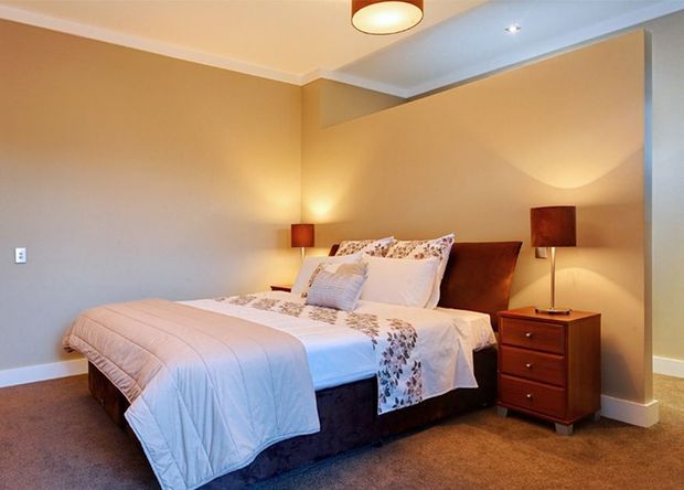A warmly lit bedroom with a large upholstered bed dressed in white and floral bedding, matching bedside lamps, and a wooden chest of drawers. The room has beige walls, beige carpet, white trim, and a simple recessed ceiling detail.