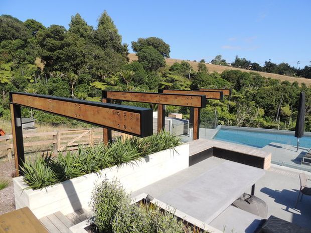 Elevated outdoor terrace overlooking a landscaped rural or semi-rural setting, with a swimming pool, paved entertaining area, built-in white planter beds, timber decking, and prominent rusted steel privacy or screening panels. The scene is sunny and contemporary, with mature vegetation and rolling fields beyond.