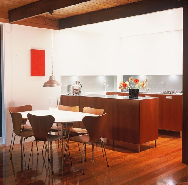 A compact mid-century-inspired kitchen and dining area with a white kitchen backdrop, dark timber ceiling beams, warm timber flooring, and a rich dark-wood kitchen island and cabinetry. A white oval dining table with sculptural wooden chairs sits in the foreground, while a stainless pendant light, reflective splashback, and bright red artwork add contrast.