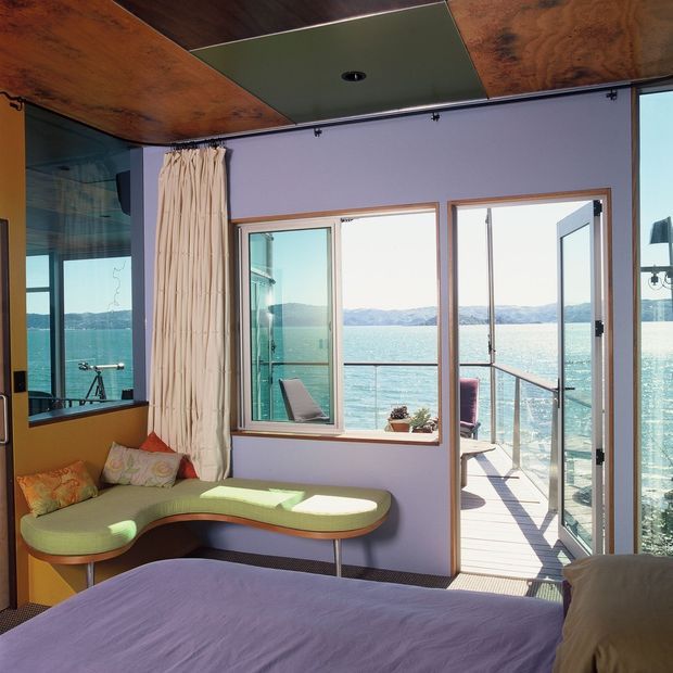 A bright waterfront bedroom or sleeping area with a bed in the foreground, a curved built-in window seat, and expansive glazing opening onto a narrow deck over turquoise water. The room features lavender-painted walls, warm wood ceiling panels, timber-framed windows and doors, and relaxed upholstered seating. Mountainous shoreline is visible beyond the water.