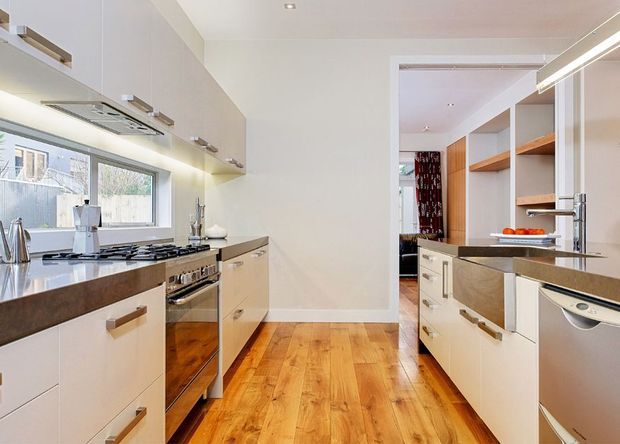 Bright contemporary galley kitchen with white flat-panel cabinetry, dark stone benchtops, stainless-steel appliances, and warm timber flooring. A large window provides natural light, while a doorway connects to additional built-in storage and shelving.