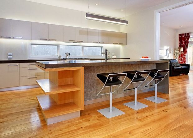 Bright contemporary open-plan kitchen with pale built-in cabinetry, stainless-steel appliances and fixtures, a dark textured tiled island face, and a timber breakfast-bar extension with open shelving. Three modern bar stools sit at the island, while timber flooring continues into an adjacent living area visible through the wide opening. Recessed ceiling lights and a suspended linear pendant provide illumination.