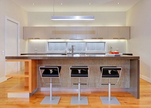 Bright contemporary kitchen with a large central island and three modern bar stools. The island has a dark stone benchtop and textured brown fixed facing, while pale wood-toned cabinetry spans the back wall beneath integrated lighting. A wide horizontal window with blinds sits above the sink, and timber flooring, recessed ceiling lights, and a suspended linear pendant complete the space.