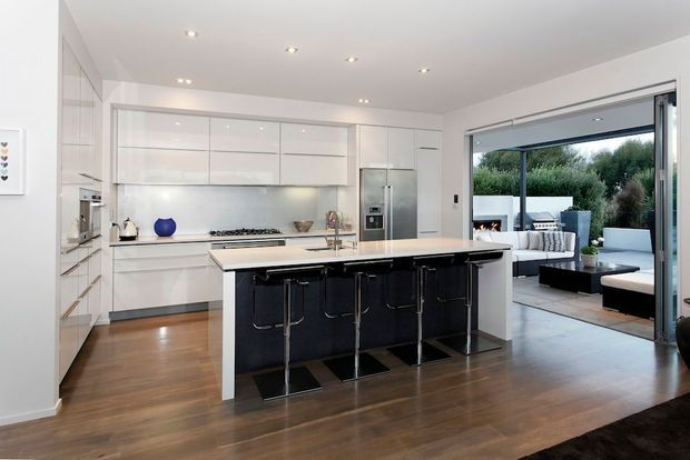 A bright, spacious contemporary open-plan kitchen with extensive white built-in cabinetry, a large central island with a dark feature base and pale benchtop, stainless-steel appliances, and four bar stools. Warm timber flooring extends into the adjoining living area, which opens through large sliding glass doors to an outdoor entertaining space. Recessed ceiling lights and clean-lined joinery create a polished, uncluttered look.