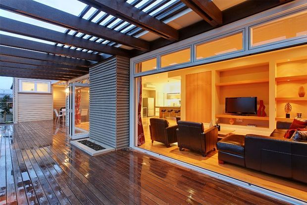 A warm contemporary outdoor entertaining deck beneath a timber pergola, looking through large glazed openings into a stylish living room. The deck has dark timber flooring and horizontal timber-clad walls, while the interior features a built-in timber entertainment unit, leather seating, and warm ambient lighting.